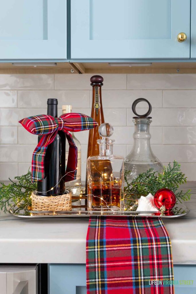Our Christmas bar top with blue cabinets and quartzite countertops, featuring a wine bottle bow topper, silver tray, plaid decanter, plaid tea towel, and twinkle lights!