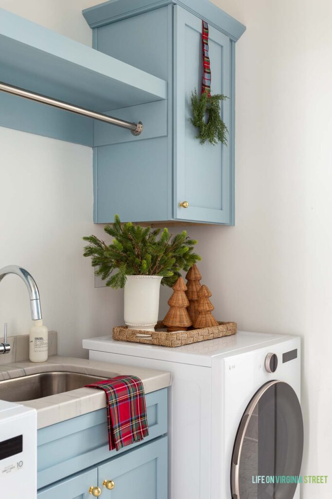 Our laundry room decorated for Christmas. I hung a mini cedar wreath with plaid ribbon on our blue cabinets, added a red Stewart plaid tea towel, vase with faux evergreens, rattan Christmas trees and a woven tray to add a festive touch!