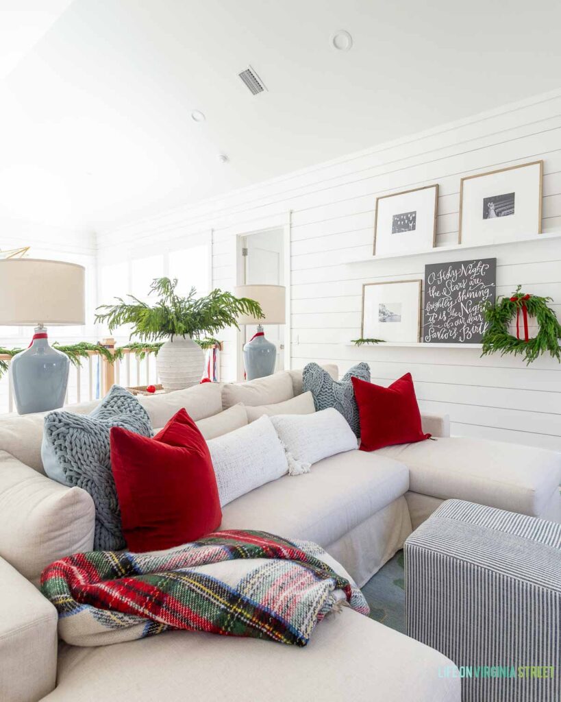 Our coastal loft with white shiplap walls decorated for Christmas with red velvet pillow and light blue knit pillows on our linen sectional, "O Holy Night" art, real-touch Norfolk pine stems in a white terra cotta vase, picture ledge gallery wall frames, and light blue ceramic lamps.