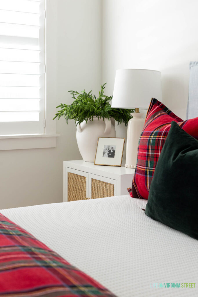 Christmas greenery stems in a white vase in a bedroom decorated with a light blue linen bed, red Stewart plaid bedding, dark green velvet pillows, a ceramic lamp, and rattan nightstand.