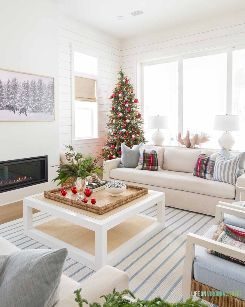 A cozy coastal Christmas living room with linen sofas, blue and white striped rug, red Stewart plaid throw pillows, a 9' Christmas tree with red and silver ornaments, evergreen stems, a raffia coffee table and cozy throw blankets.