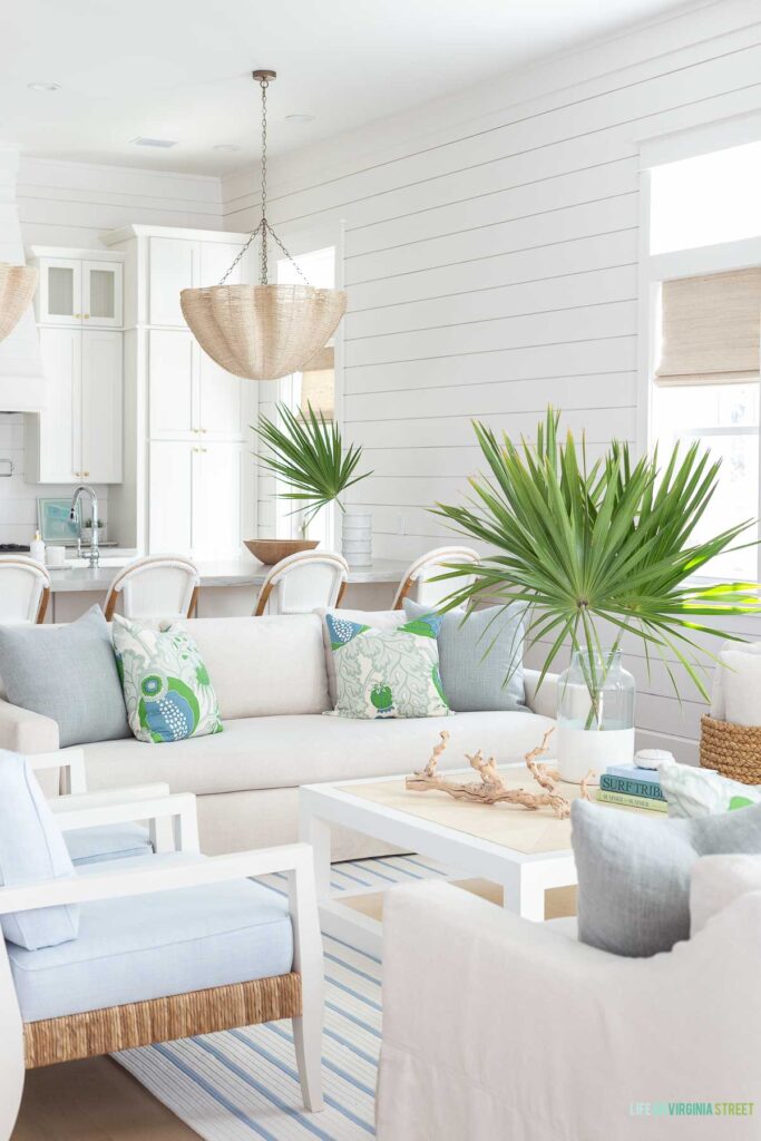 A coastal living room with linen sofas, blue and white striped rug, raffia coffee table and rope chandelier. Accented with blue and green decor. The shiplap walls are painted Sherwin Williams Snowbound.
