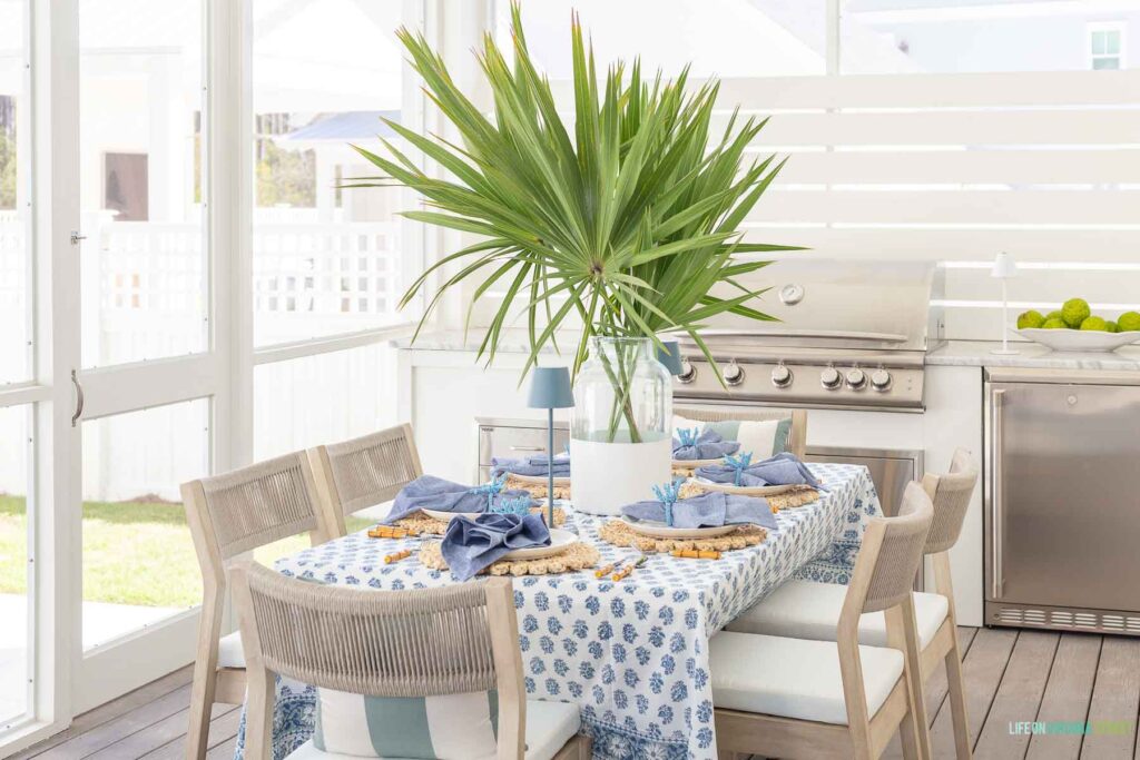 A coastal outdoor kitchen and dining space with rope dining chairs, a blue block print tablecloth, large palm fronds, blue linen napkins and built-in grill and outdoor fridge.