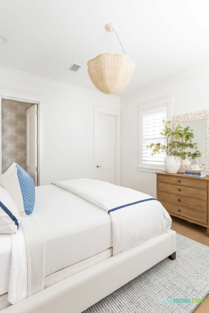 A coastal guest bedroom featuring a white upholstered bed, luxe bedding, a seashell mirror, blue grid rug, scalloped wicker chandelier, palm print wallpaper, and a light wood dresser.