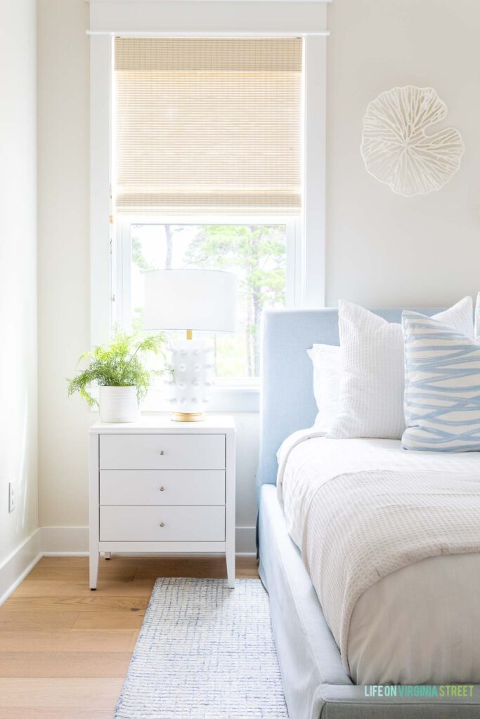 A coastal bedroom painted Sherwin Williams White Duck, one of the best coastal paint colors! This beach house bedroom also features woven roman shades, a light blue upholstered bed, blue and white bed linens, a white nightstand, and white oak hardwood floors.