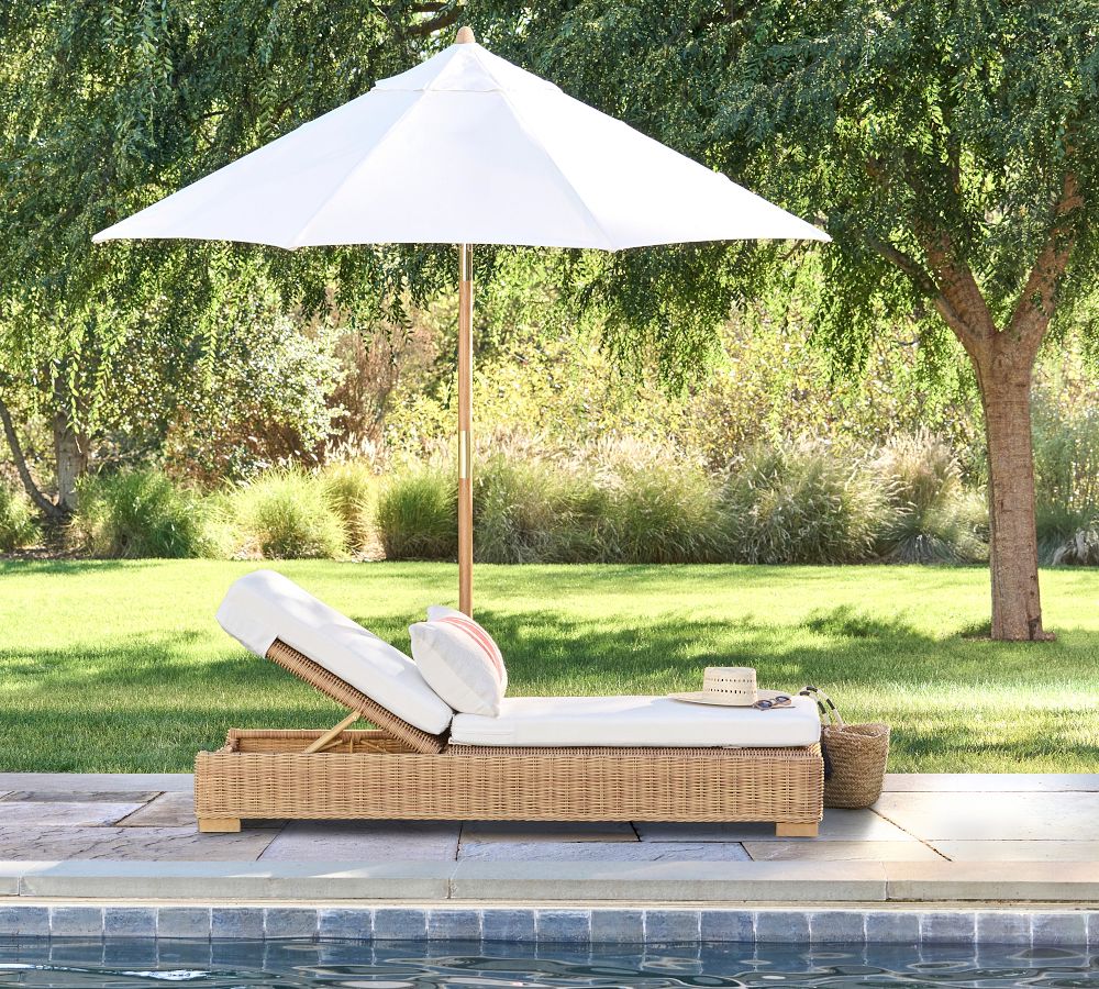 Natural wicker colored pool lounge chair with a white outdoor cushion and nearby white umbrella near a swimming pool.