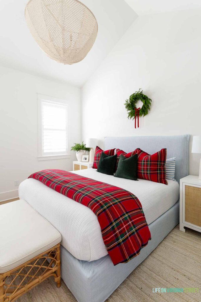 Christmas bedroom with vaulted ceilings, a light blue linen bed, white and red plaid bedding, dark green velvet pillows, a wreath with red velvet bow above the bed, bead chandelier, rattan nightstands, a rattan bench, and white jute rug.