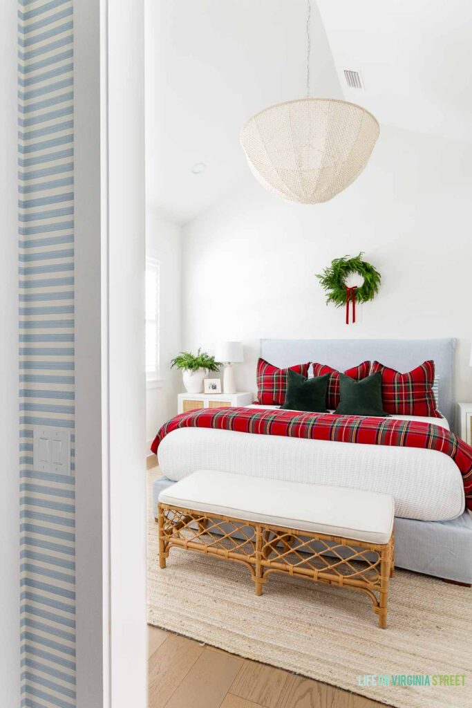 Christmas bedroom with a light blue linen bedframe, red Stewart plaid bedding, dark green velvet pillows, a wreath with red velvet bow above the bed, bead chandelier, rattan nightstands, a rattan bench, and white jute rug.