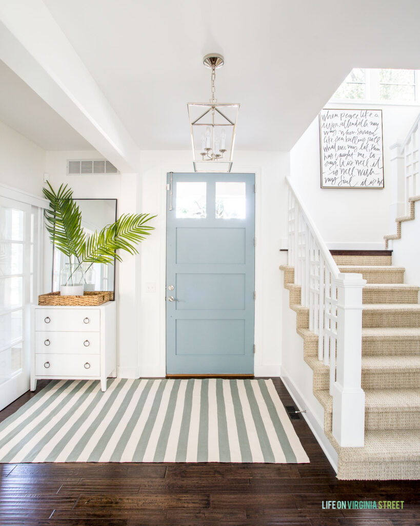 The inside of a front door painted Benjamin Moore Water's edge, one of the best coastal paint colors. Includes a white raffia cabinet, chrome lantern pendant light, palm fronds, and blue striped rug.